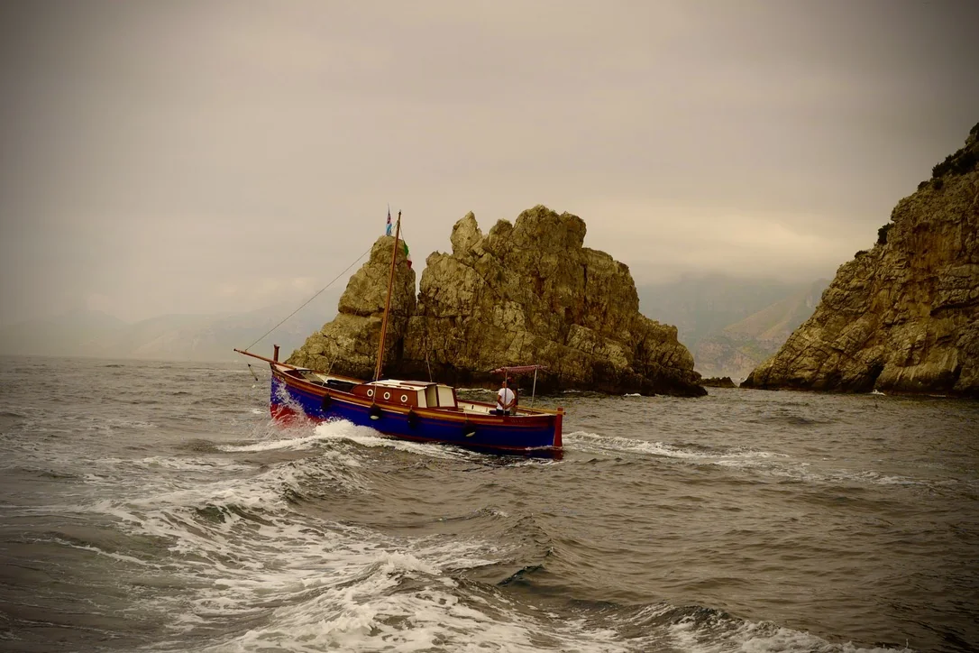 A small red and blue boat crossing choppy water between rocky shores